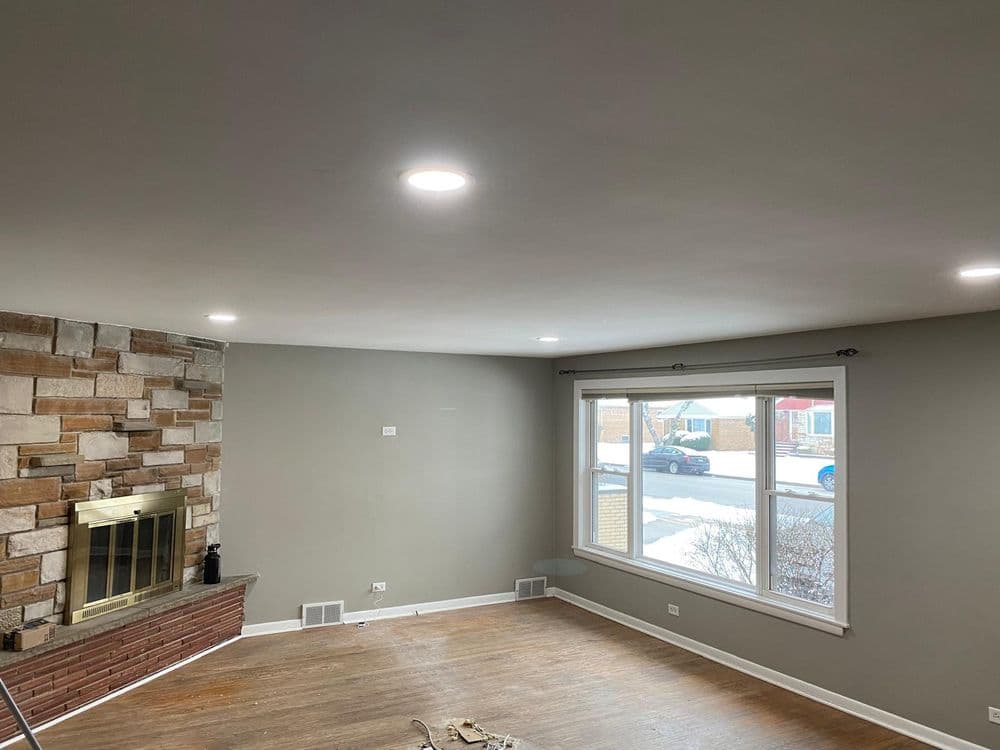 Modern living room with stone fireplace and large window, featuring recessed lighting and hardwood floors.