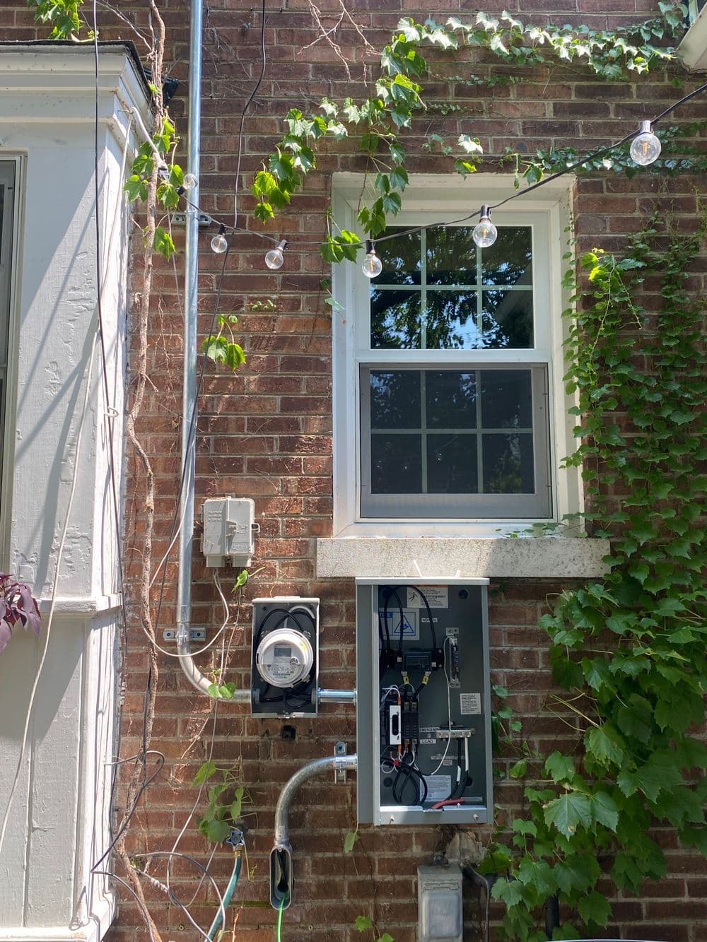 Electric meter and service panel on a brick wall with ivy and string lights outdoors.