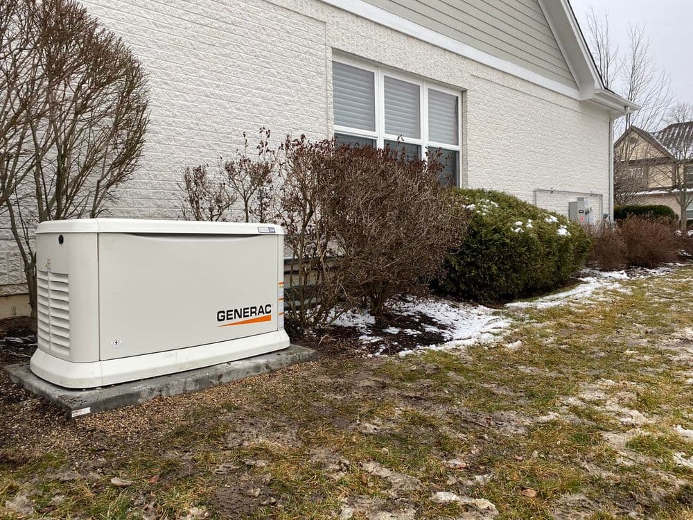 Generac home standby generator beside a house with winter landscape and shrubs.