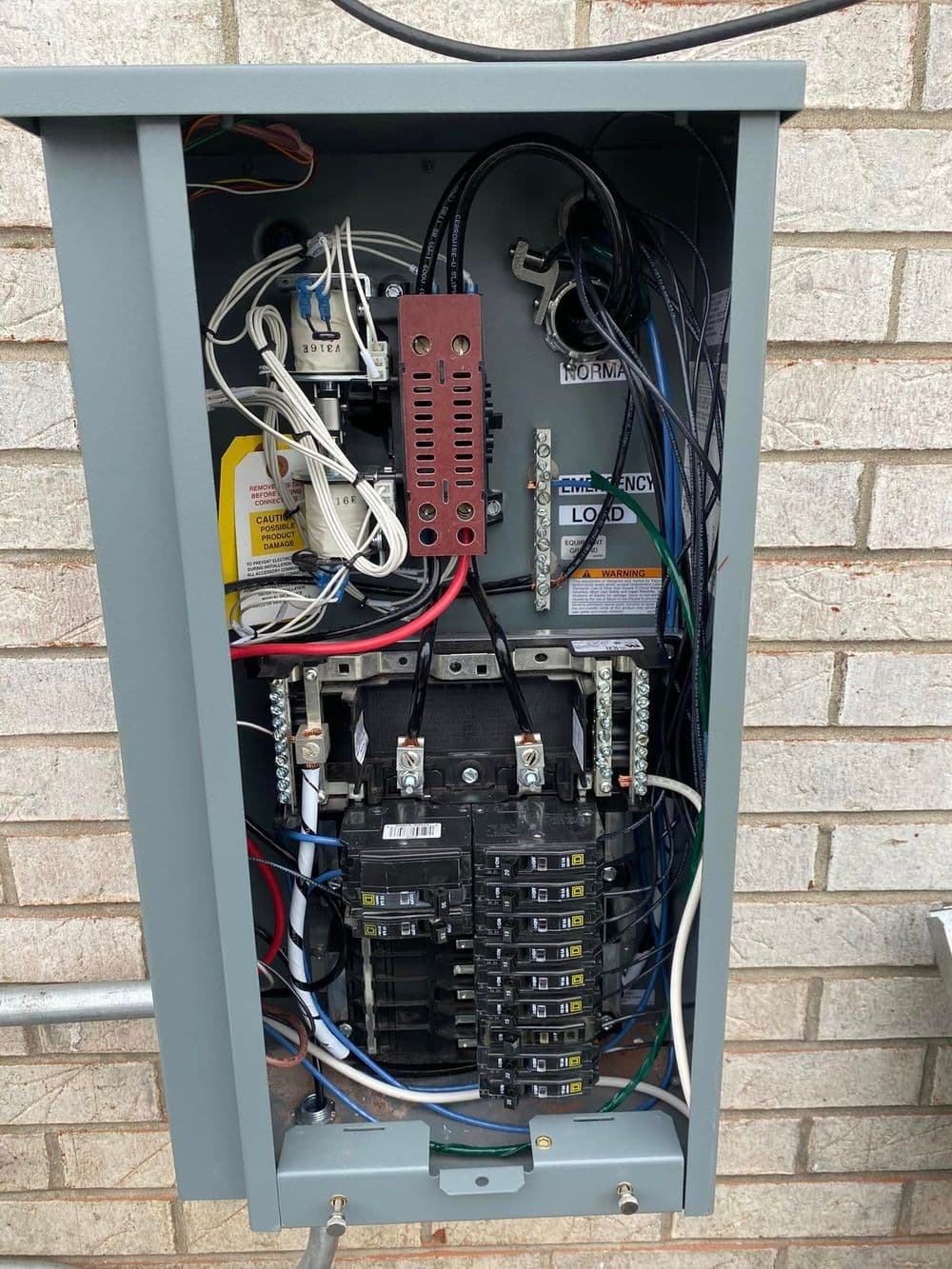 Electrical panel with circuit breakers and wiring connections mounted on brick wall.